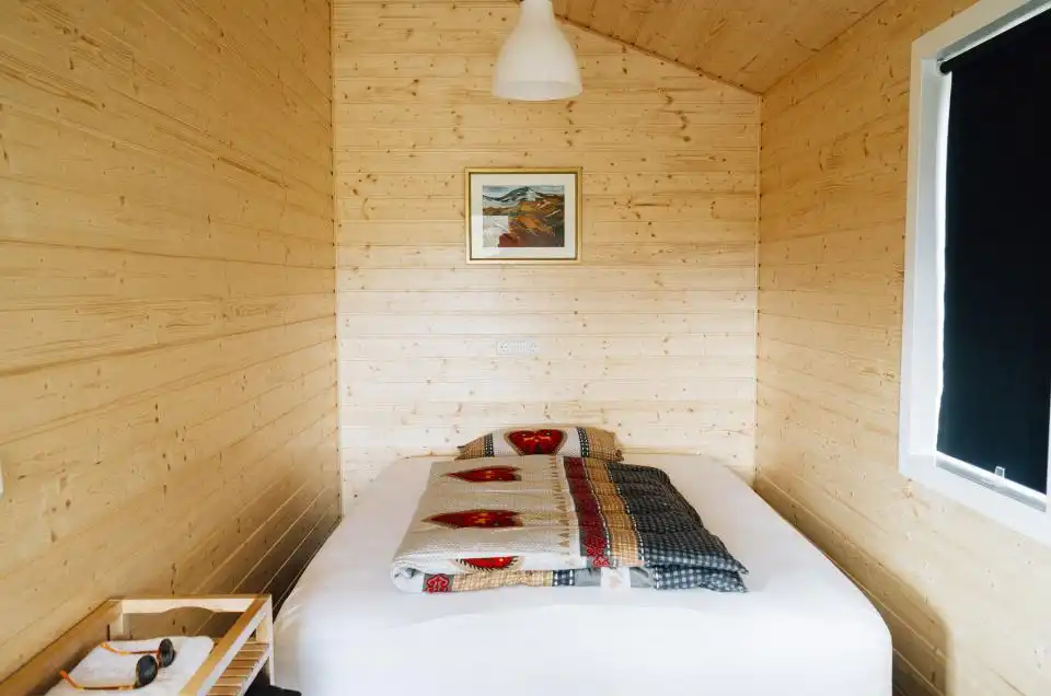 Quarto revestido de madeira clara, com cama arrumada e cortina escura